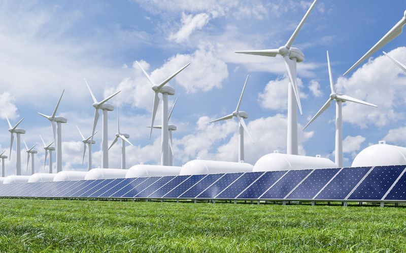 Una granja de energía renovable con hileras de paneles solares en primer plano y múltiples turbinas eólicas bajo un cielo nublado.