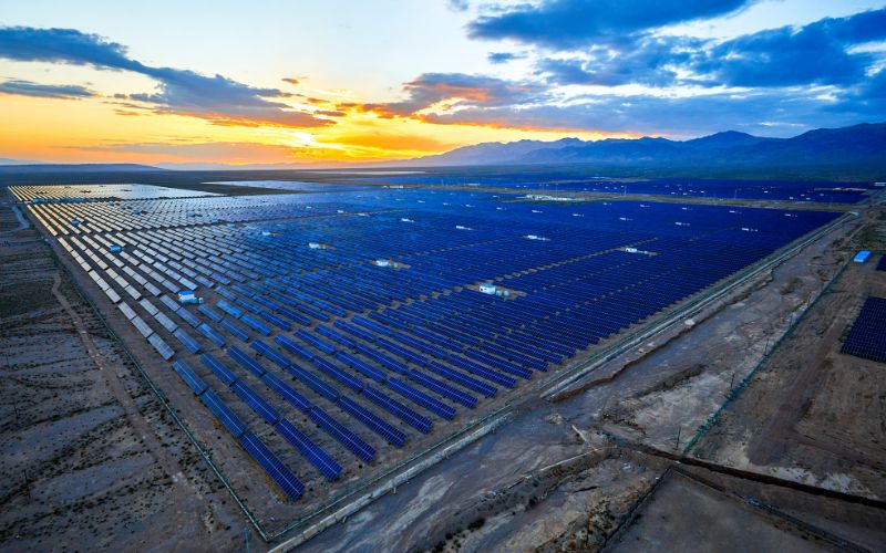 Luftaufnahme einer großen Solarfarm bei Sonnenuntergang mit Reihen von Photovoltaikmodulen, die über eine weite Landschaft vor einer Bergkulisse verteilt sind.