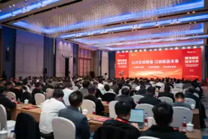 Bright, modern conference room filled with a large audience attending a corporate event with a red digital display and elegant lighting.